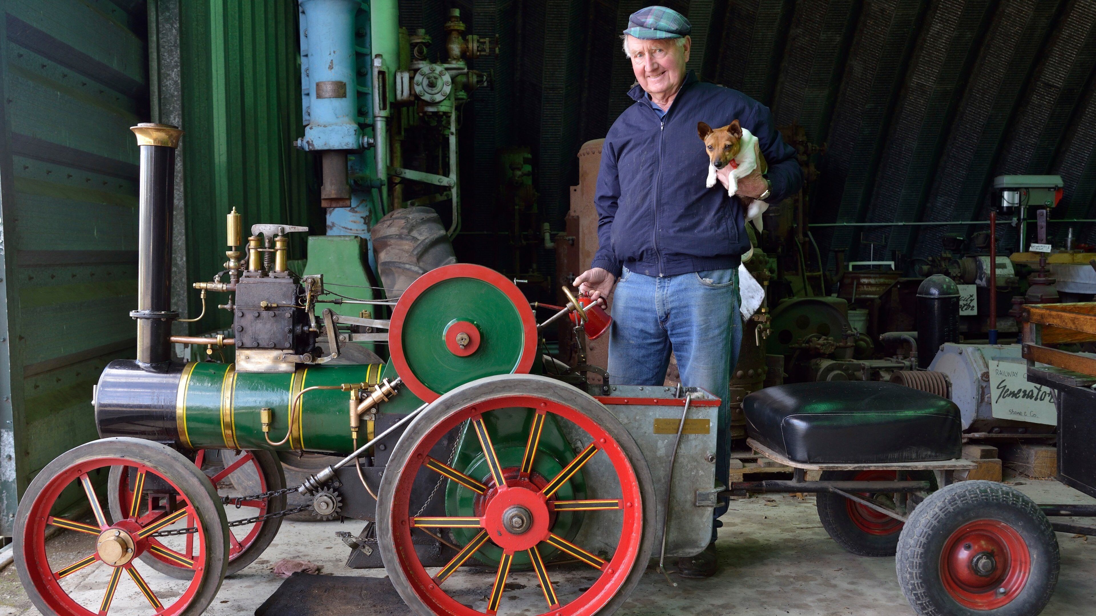 A man holding a small dog next to a vintage steam engine with large red wheels.
