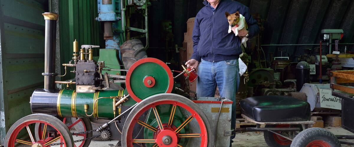 A man holding a small dog next to a vintage steam engine with large red wheels.