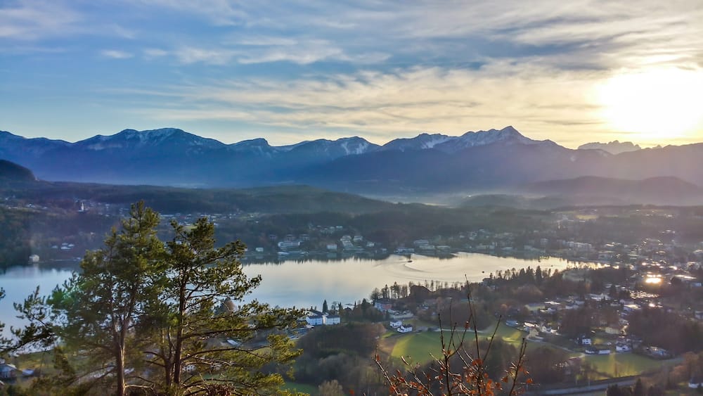 A panoramic view on an alpine landscape of Austria. There is a lake in the middle, surrounded by high mountains. Sun is slowly setting behind the horizon. Few households around the lake