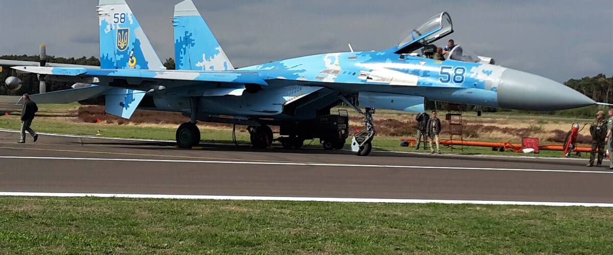 A blue fighter jet with the number 58 on its nose and tail, parked on a runway with grassy areas and a cloudy sky in the background.