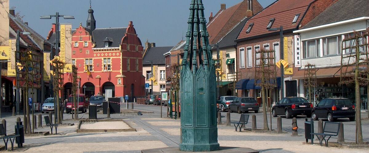 A square with a green monument, historic buildings, and parked cars.