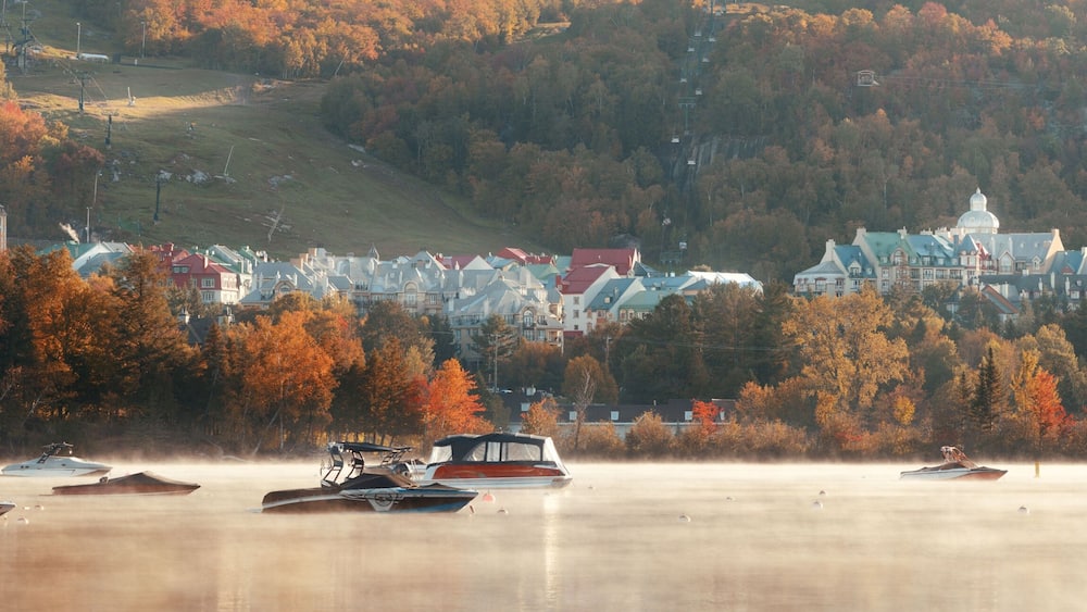 Une ville au bord d’un lac, où l’on peut admirer les couleurs automnales des arbres, des bateaux et une église perchée sur une colline.