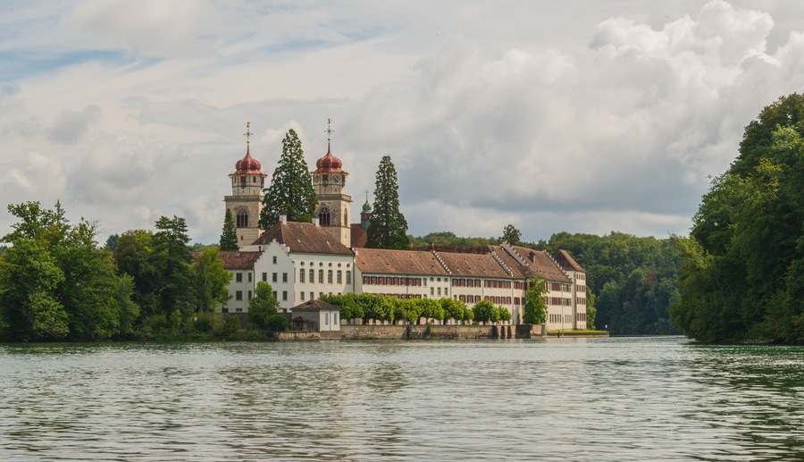 Historic castle situated by a serene lake under a cloudy sky surrounded by lush greenery