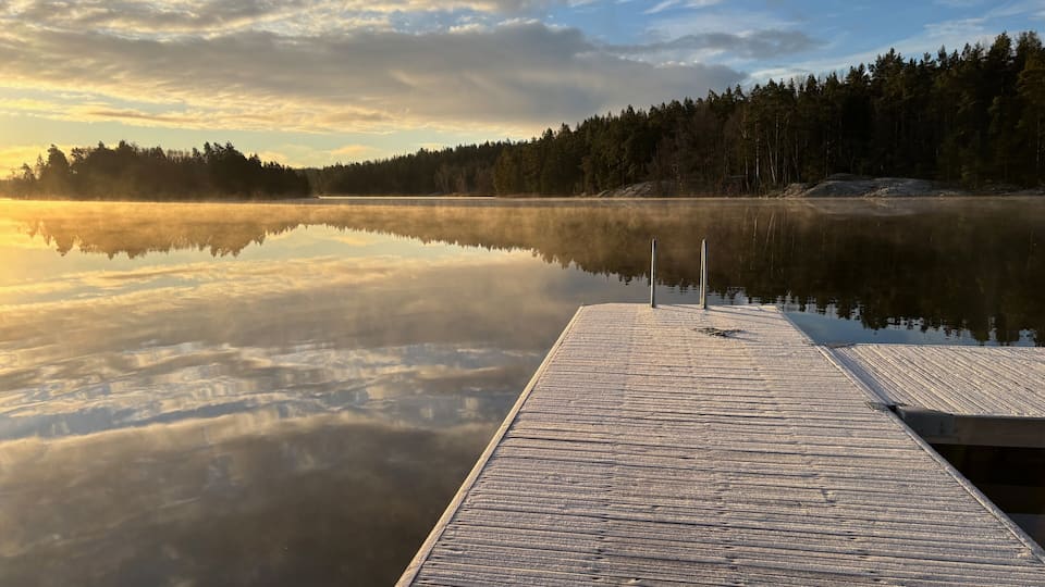 A wooden dock extending into calm waters with a forested shoreline and a cloudy sky.