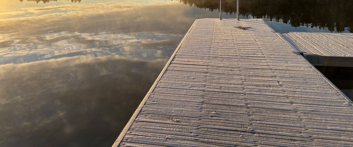 A wooden dock extending into calm waters with a forested shoreline and a cloudy sky.