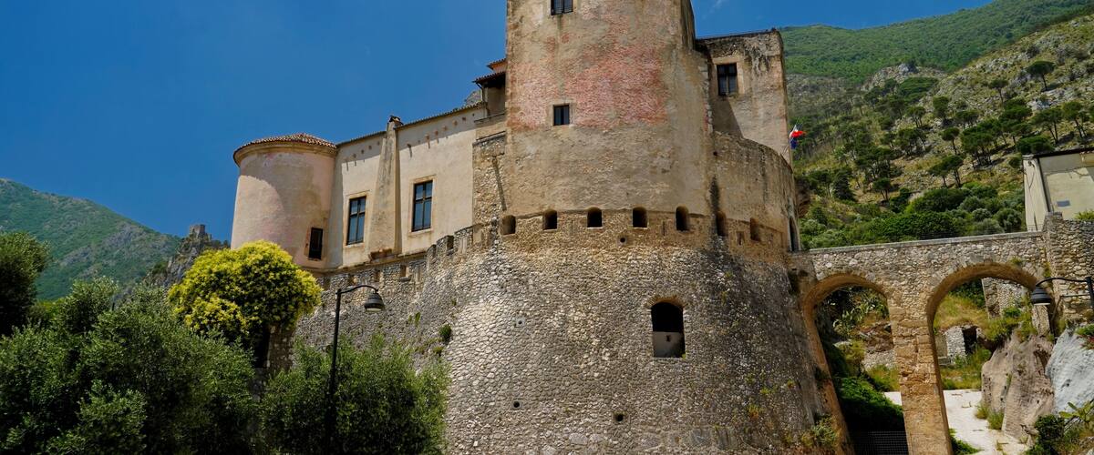 A historic stone castle with a round tower, surrounded by lush greenery and mountains.