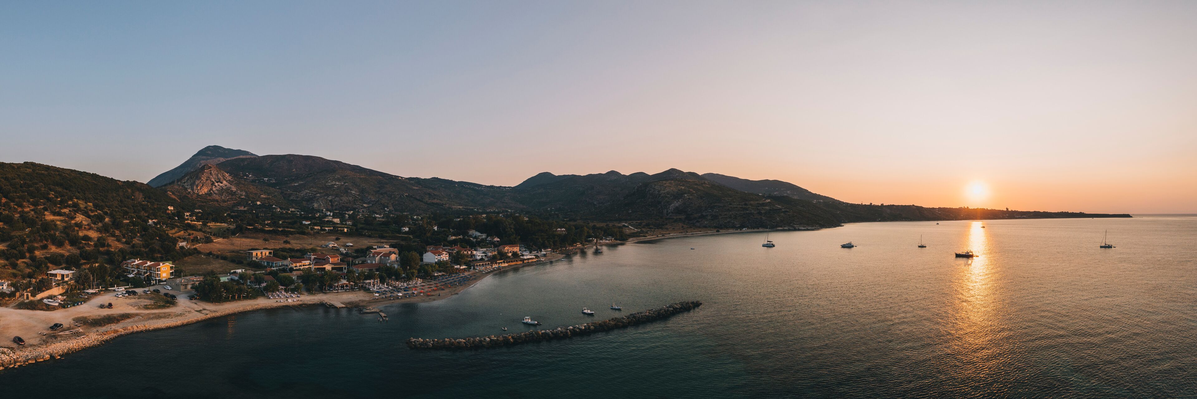 A coastal town at sunset with boats on the water and mountains in the background.