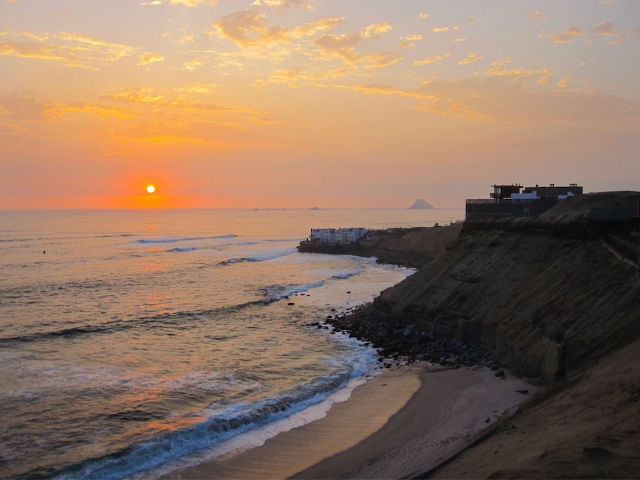 Un atardecer sobre una playa con olas, un acantilado rocoso y una isla distante.