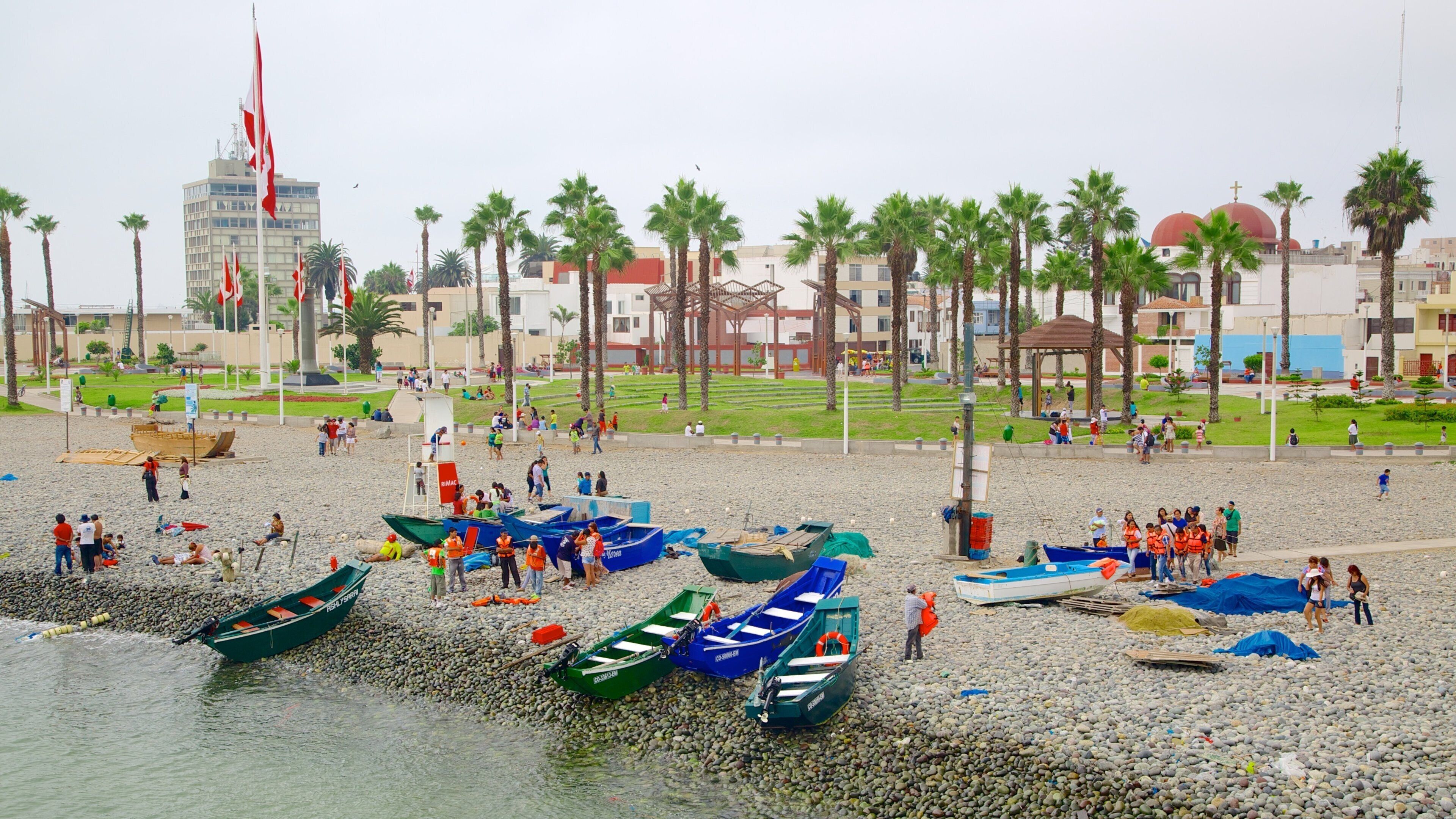 A pebbled beach with several boats, palm trees, and a building with a red dome in the background.