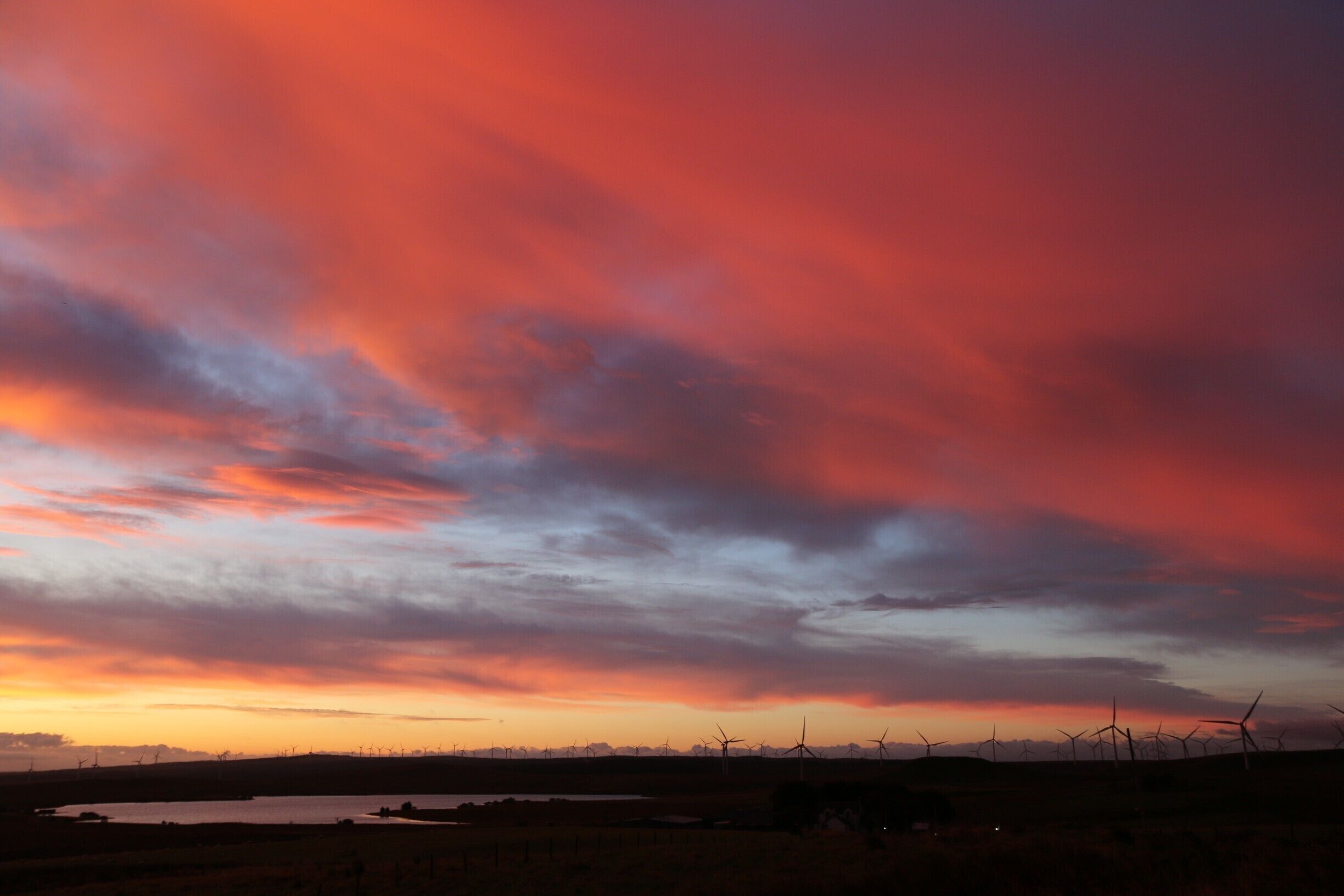 A sunset over a wind farm with a body of water in the foreground.