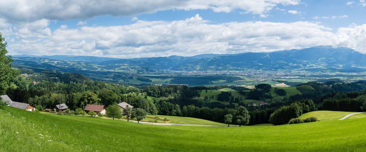 A lush green valley with rolling hills, a winding road, and a distant mountain range under a partly cloudy sky.