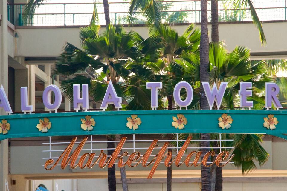 Een bord met de tekst 'ALOHA TOWER Market Place', met palmbomen op de achtergrond.