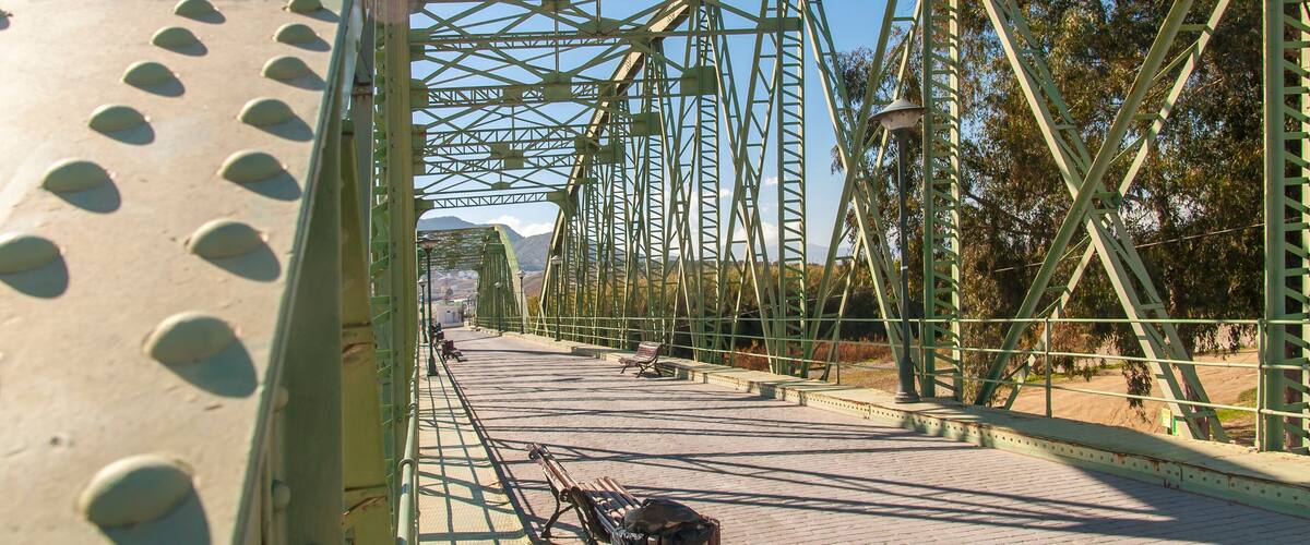 A green metal bridge with riveted beams and a paved walkway.