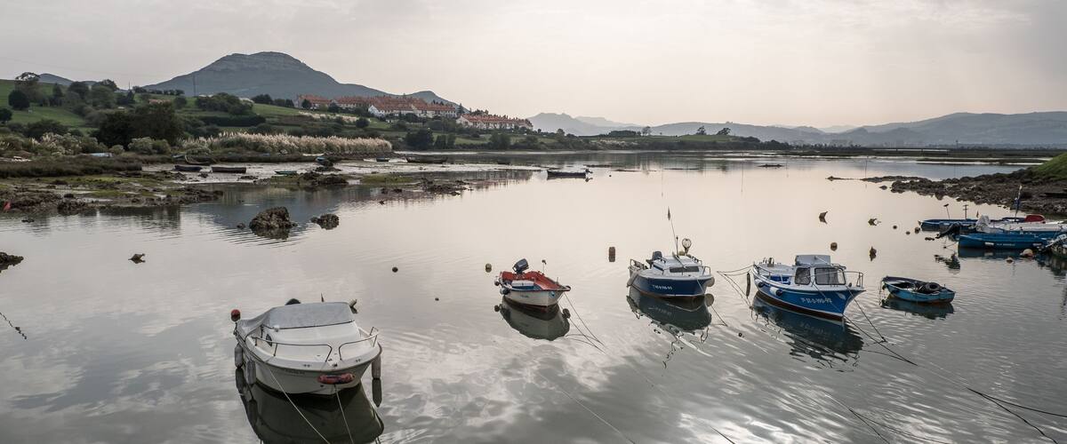 A calm lake with several boats anchored, surrounded by hills and a distant mountain range.