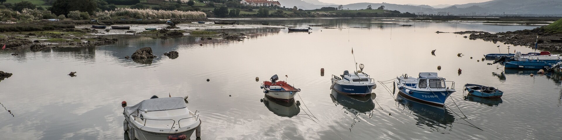 A calm lake with several boats anchored, surrounded by hills and a distant mountain range.