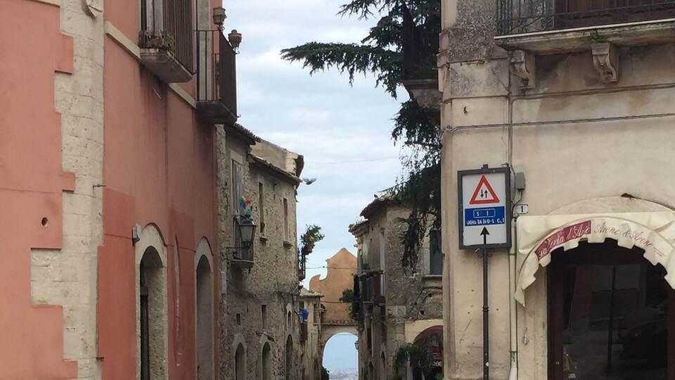 A cobblestone street flanked by historic buildings with arched windows and balconies. A triangular warning sign is visible on the right.