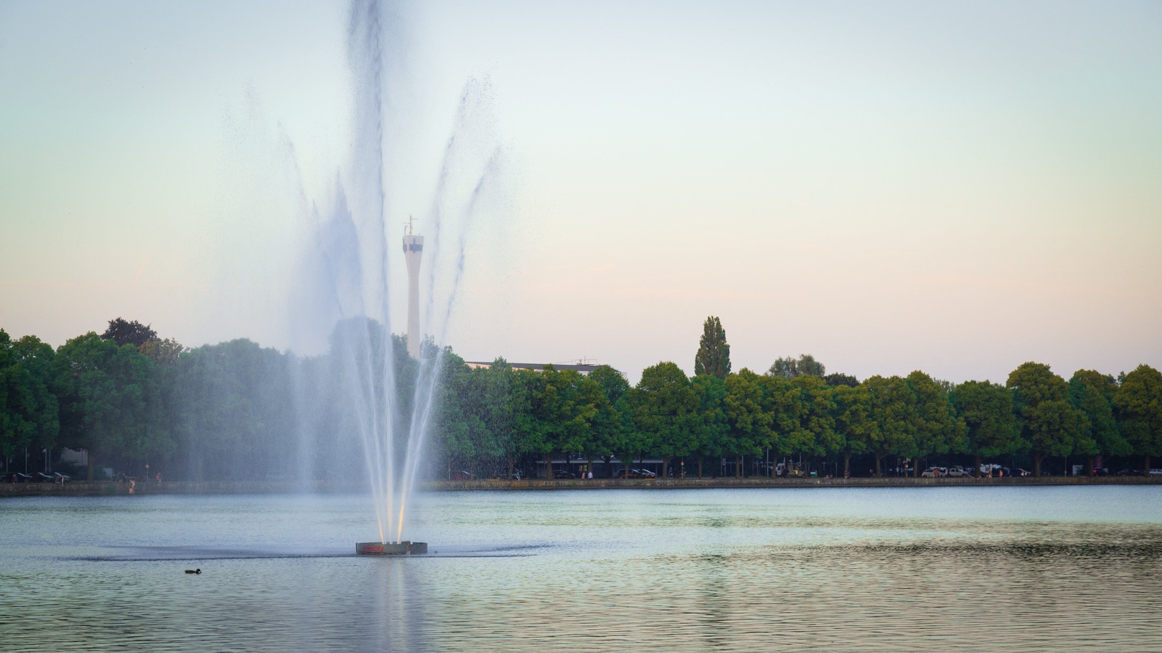 Une fontaine dans un lac, avec des arbres et une tour en arrière-plan.
