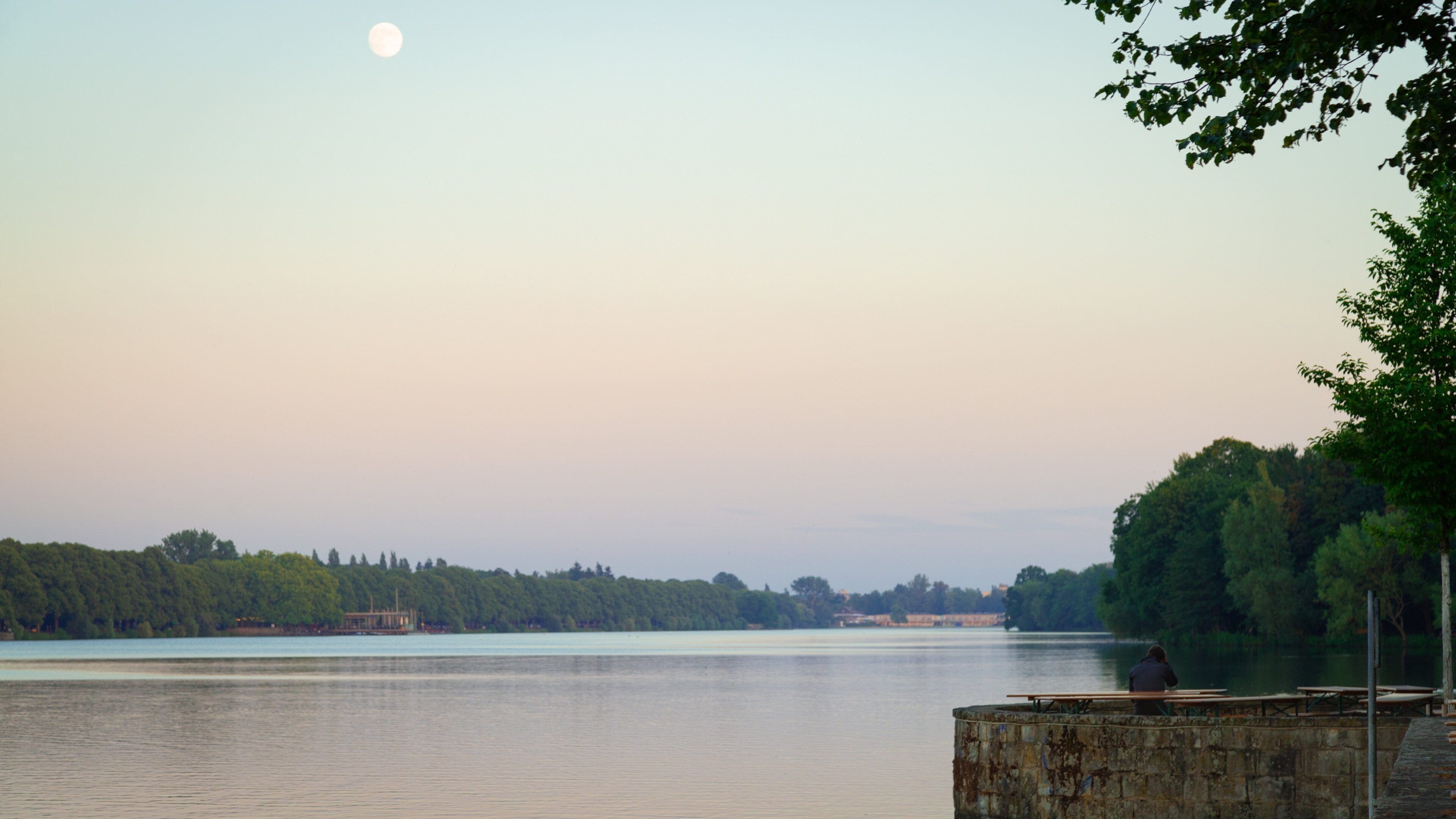 A calm lake with a wooden pier and picnic tables, surrounded by trees and a distant shoreline.