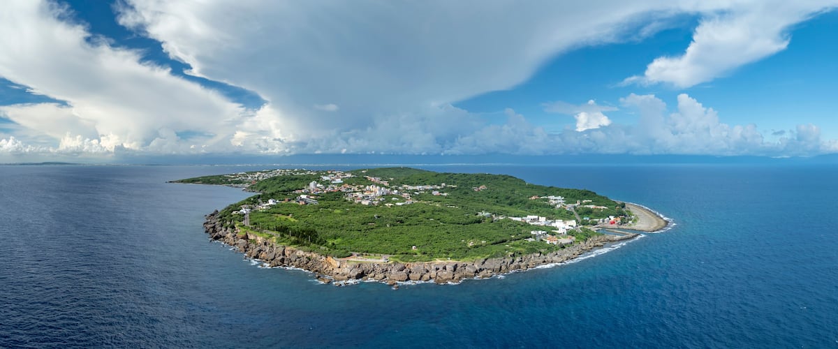 An island with a coastline, surrounded by a large body of water, under a partly cloudy sky.