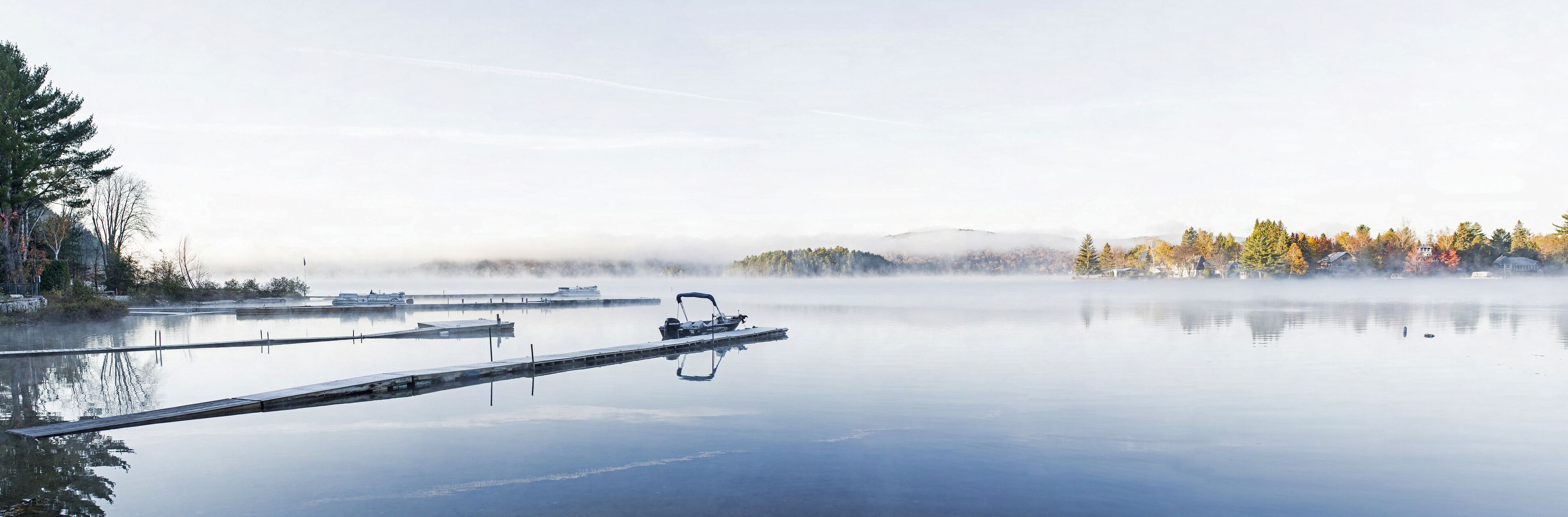 A calm lake with a dock and a boat, surrounded by autumn trees and mist-covered hills.