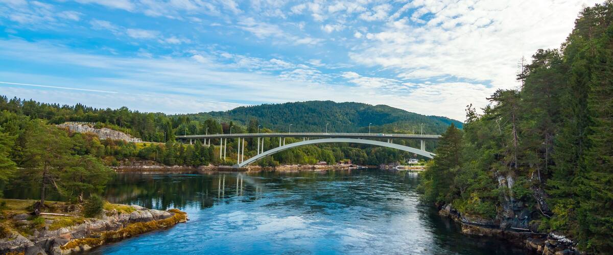 A river with a white bridge, surrounded by greenery and hills.