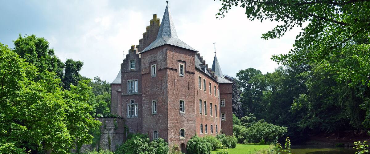 A historic brick building with a tower, surrounded by lush greenery and a calm pond.
