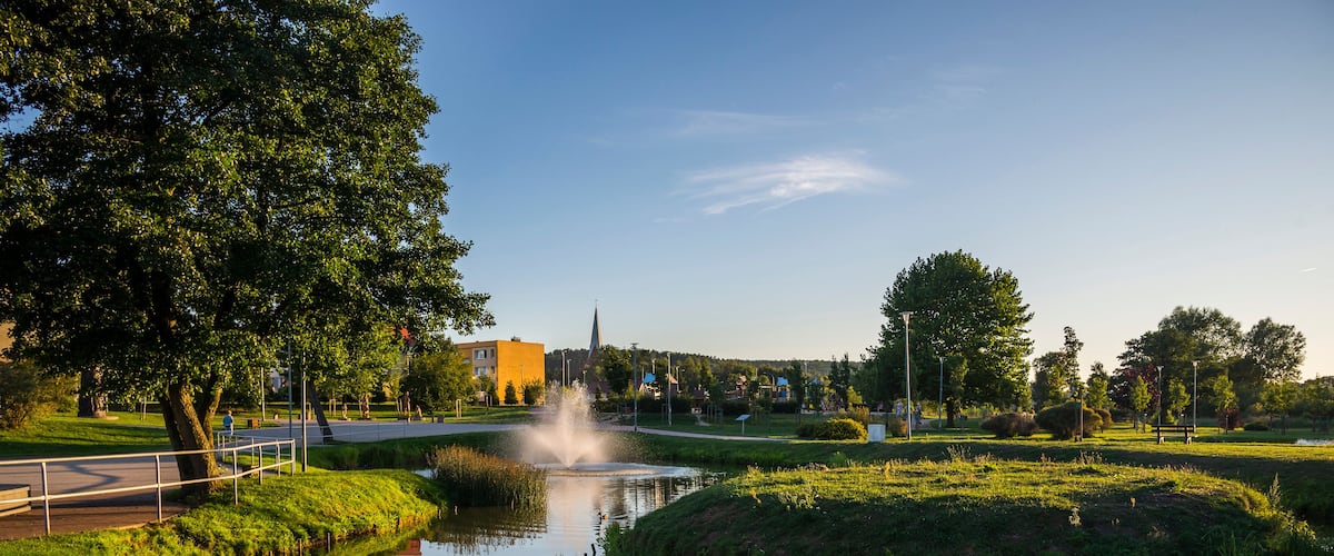 A calm river with a fountain, surrounded by lush greenery and a distant church spire.