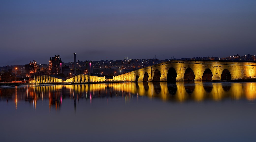 Un puente al anochecer, con sus luces reflejadas en el agua y un horizonte urbano al fondo, destacándose una torre imponente.