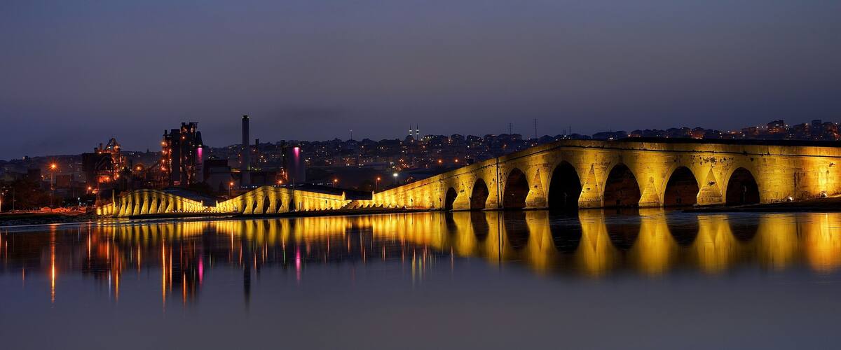 Eine Brücke im Dämmerlicht mit Lichtern, die sich im Wasser spiegeln, eine Stadtansicht im Hintergrund und ein markanter Turm.