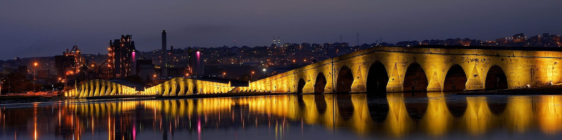A bridge at dusk with its lights reflecting on the water, a city skyline in the background, and a prominent tower.