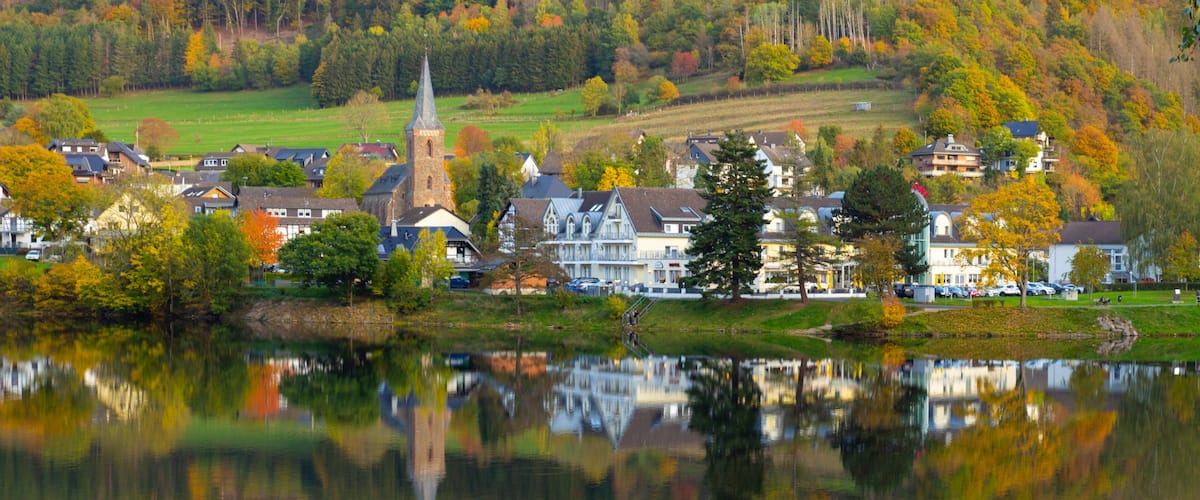 A lakeside town with a church steeple, houses, and autumn-colored trees.