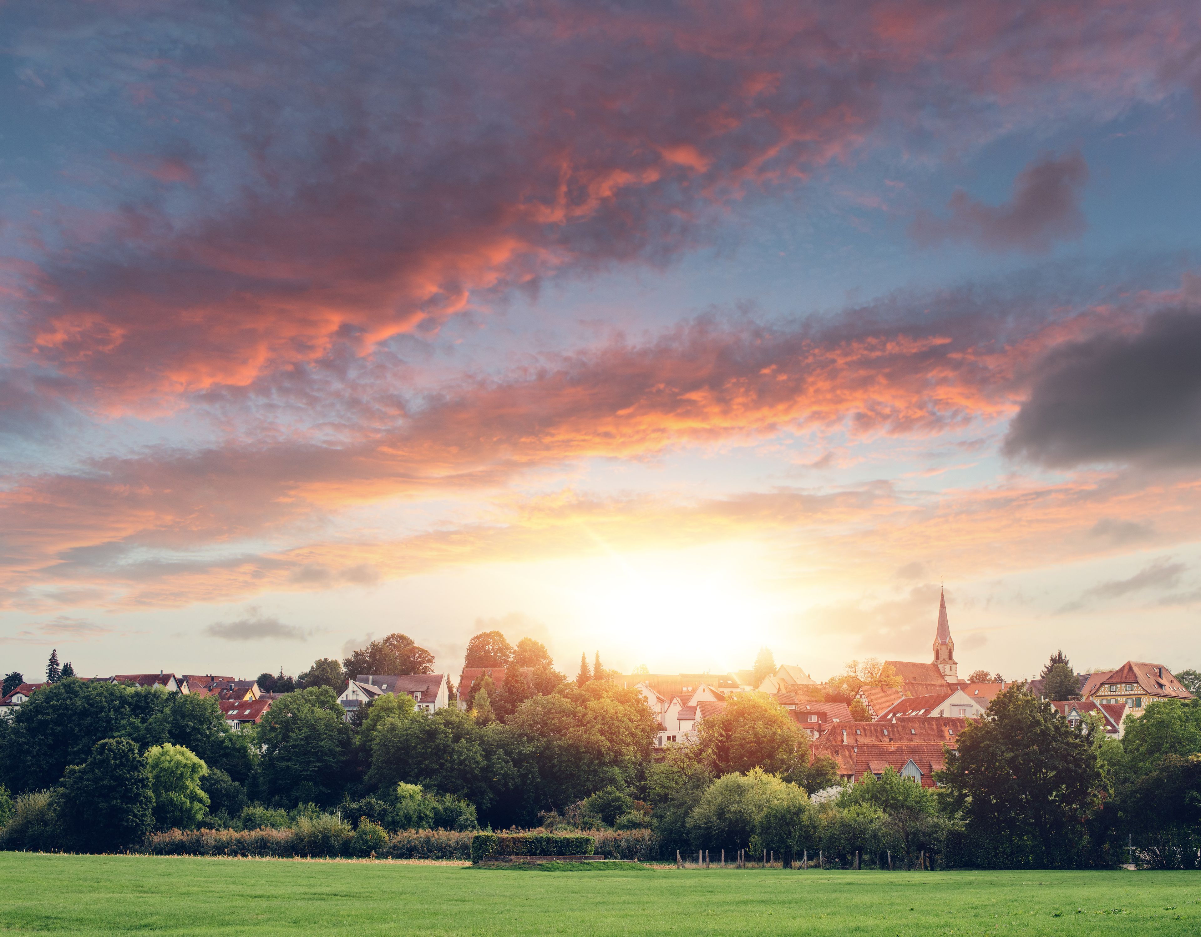 Un coucher de soleil sur une ville, avec le clocher d’une église imposante qui la domine, le tout entouré d’une végétation luxuriante et de bâtiments résidentiels.