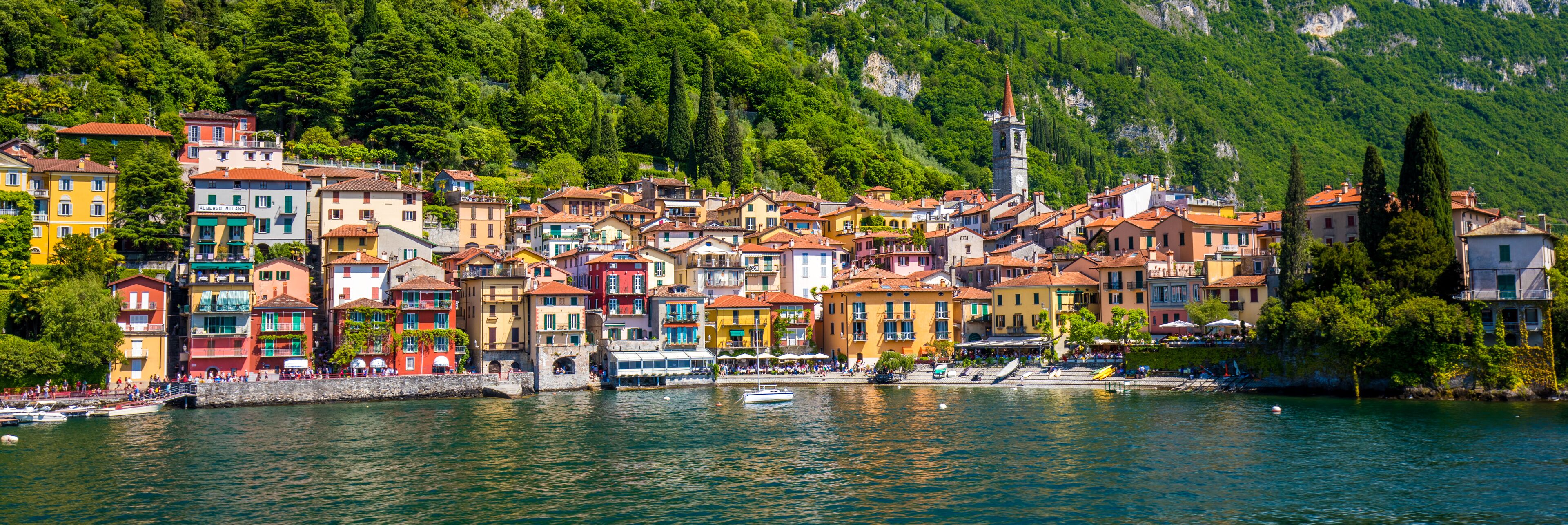 A colorful coastal town with buildings along the waterfront, a church tower, and lush green hills in the background.