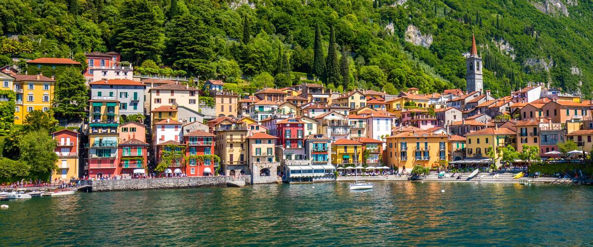 A colorful coastal town with buildings along the waterfront, a church tower, and lush green hills in the background.