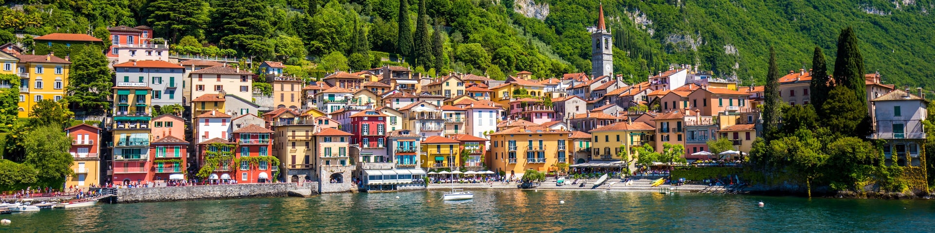 A colorful coastal town with buildings along the waterfront, a church tower, and lush green hills in the background.