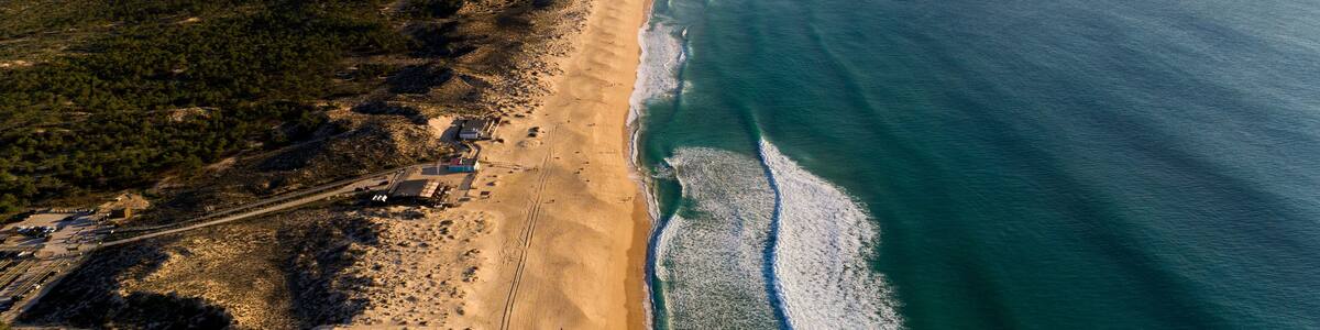 An aerial view of a coastal area with a sandy beach, a forested area, and a clear blue ocean.