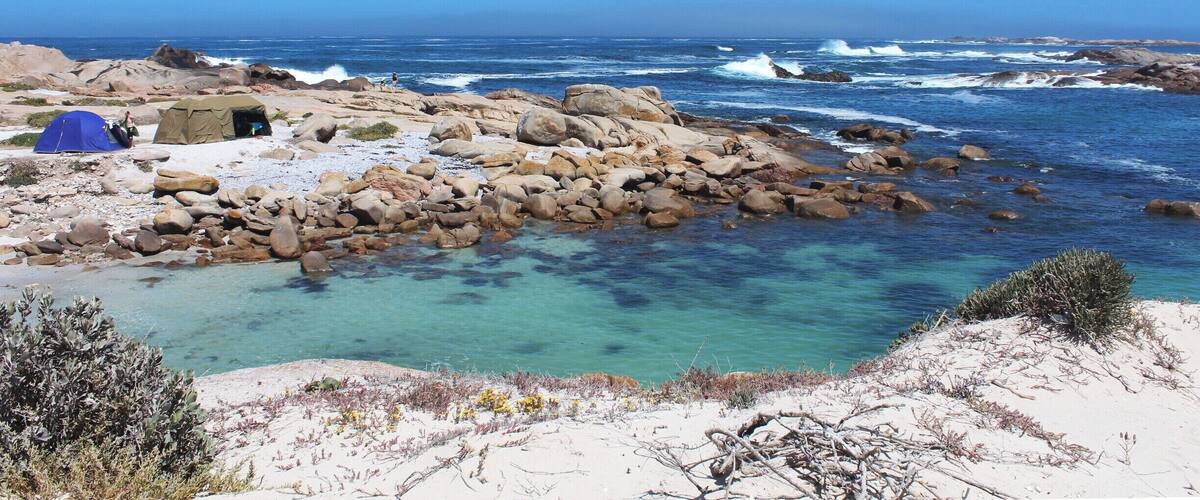 A rocky beach with clear turquoise water, a blue tent, and a rocky shoreline.