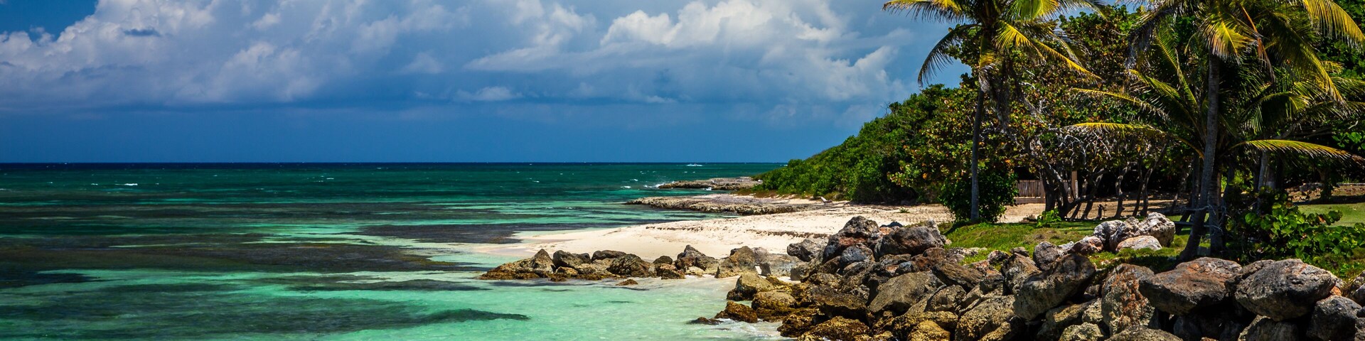 Uma praia tropical com águas turquesa cristalinas, areia branca e palmeiras, sob grandes nuvens.