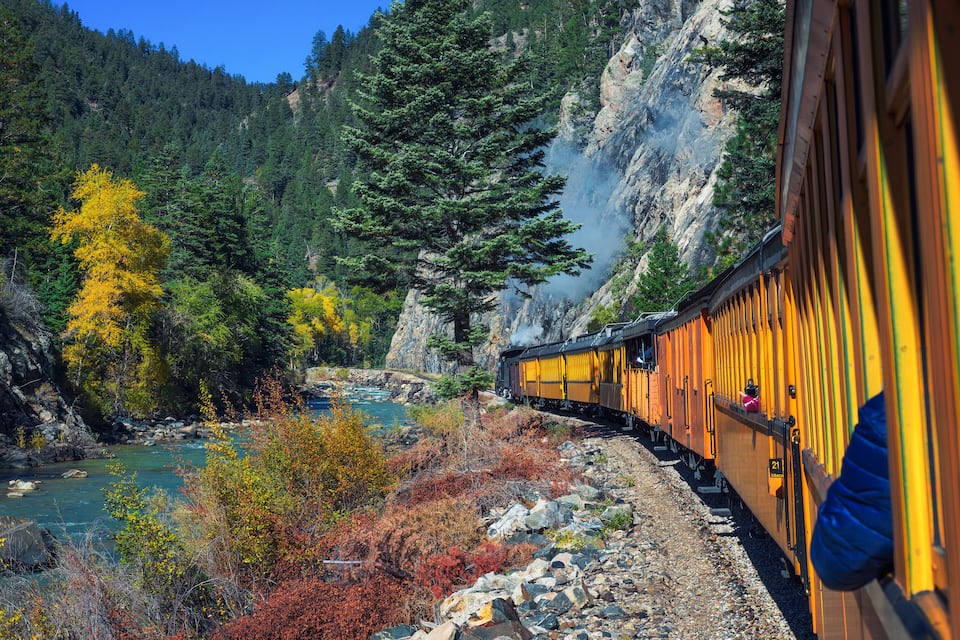 Um trem com vagões amarelos atravessando uma região montanhosa com um rio e árvores com folhagem avermelhada e amarelada.