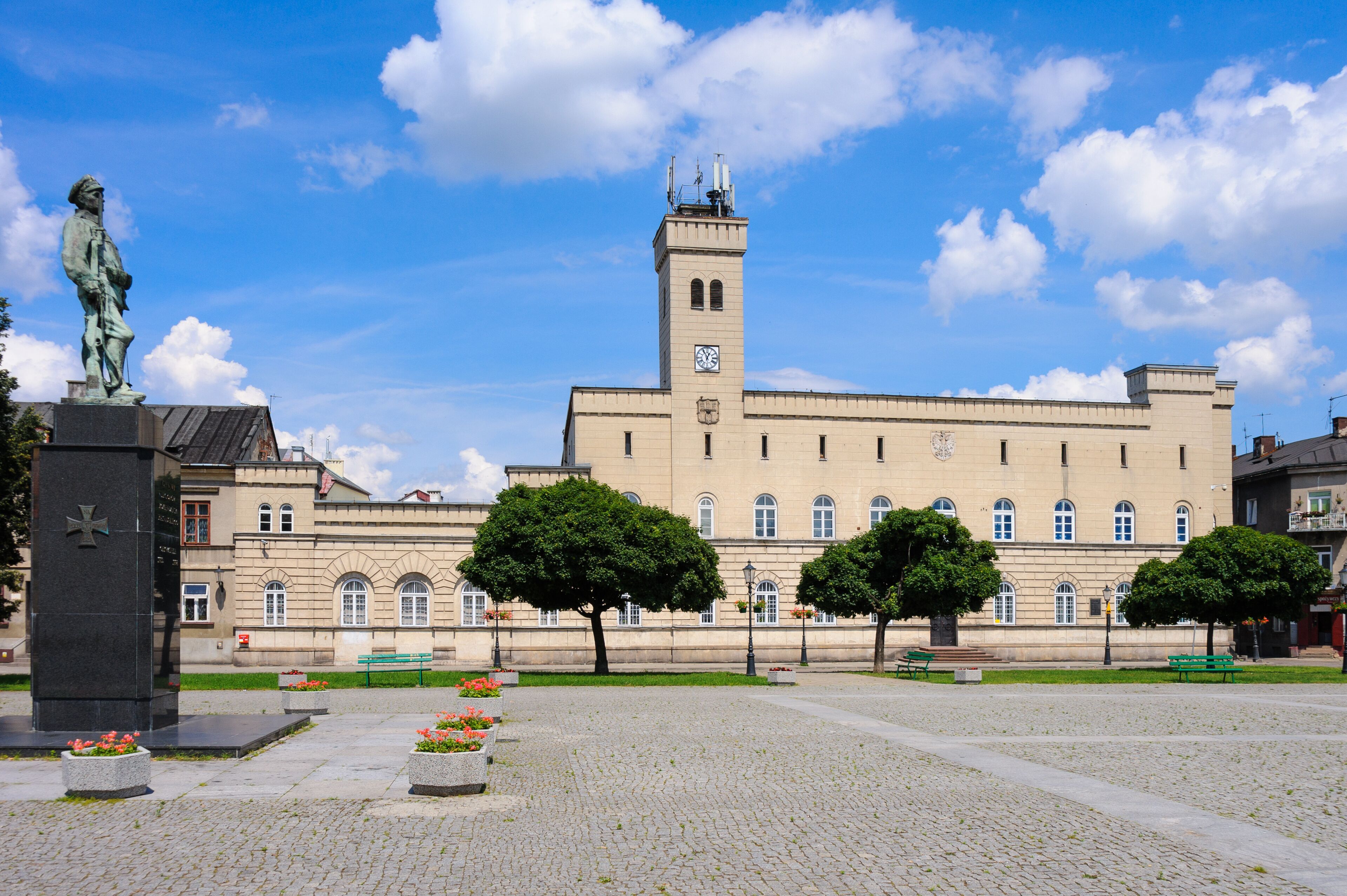 A historic building with a clock tower, a statue, and a cobblestone plaza with benches and trees.