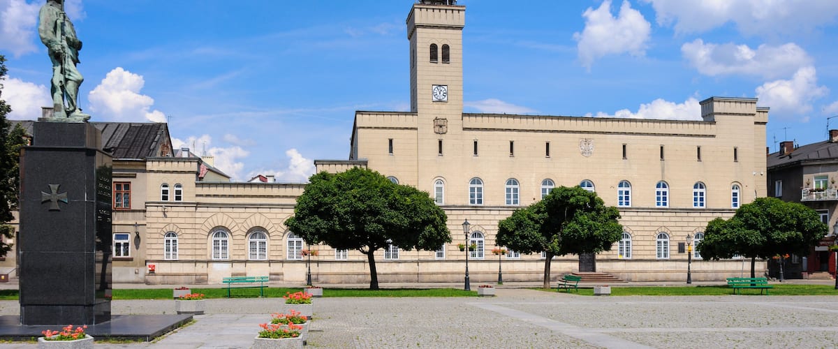 A historic building with a clock tower, a statue, and a cobblestone plaza with benches and trees.