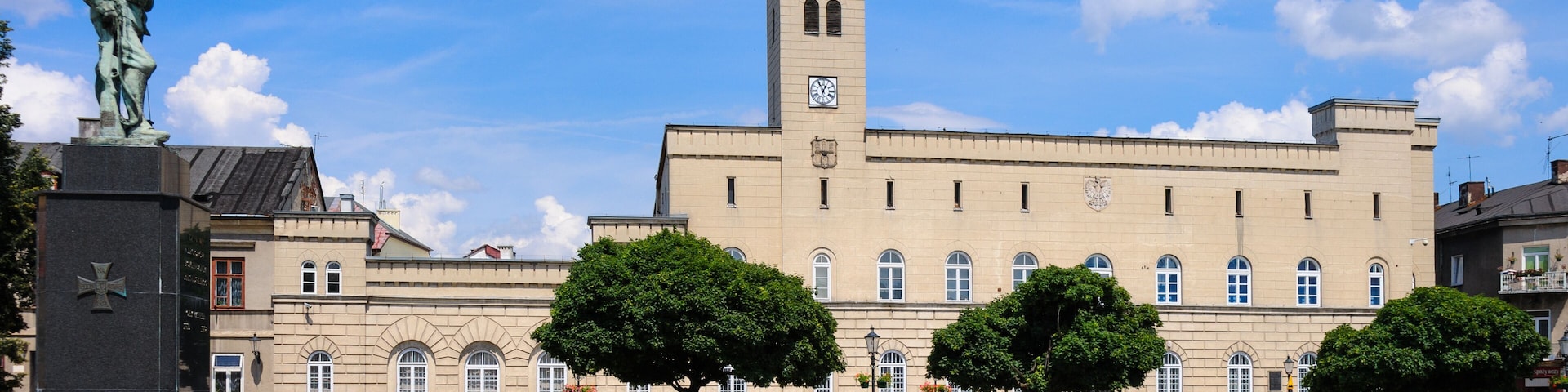 A historic building with a clock tower, a statue, and a cobblestone plaza with benches and trees.