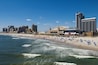 A coastal cityscape with a beach, ocean waves, and various buildings including a tall hotel and a stadium.