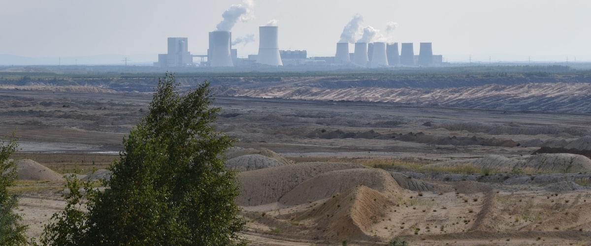 A power plant with multiple cooling towers emitting steam, surrounded by a flat, barren landscape with sparse vegetation.
