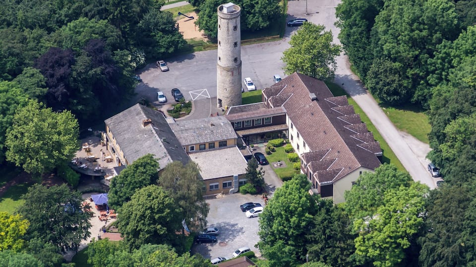 An aerial view of a historic building complex with a prominent tower, surrounded by lush greenery and a parking area.
