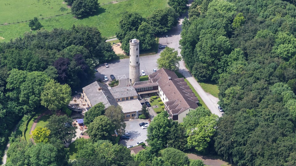 An aerial view of a historic building with a tower, surrounded by lush greenery and a parking area.