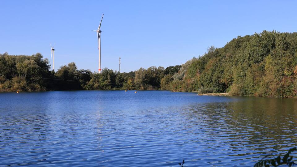 A calm lake with wind turbines in the background and a forested hillside.
