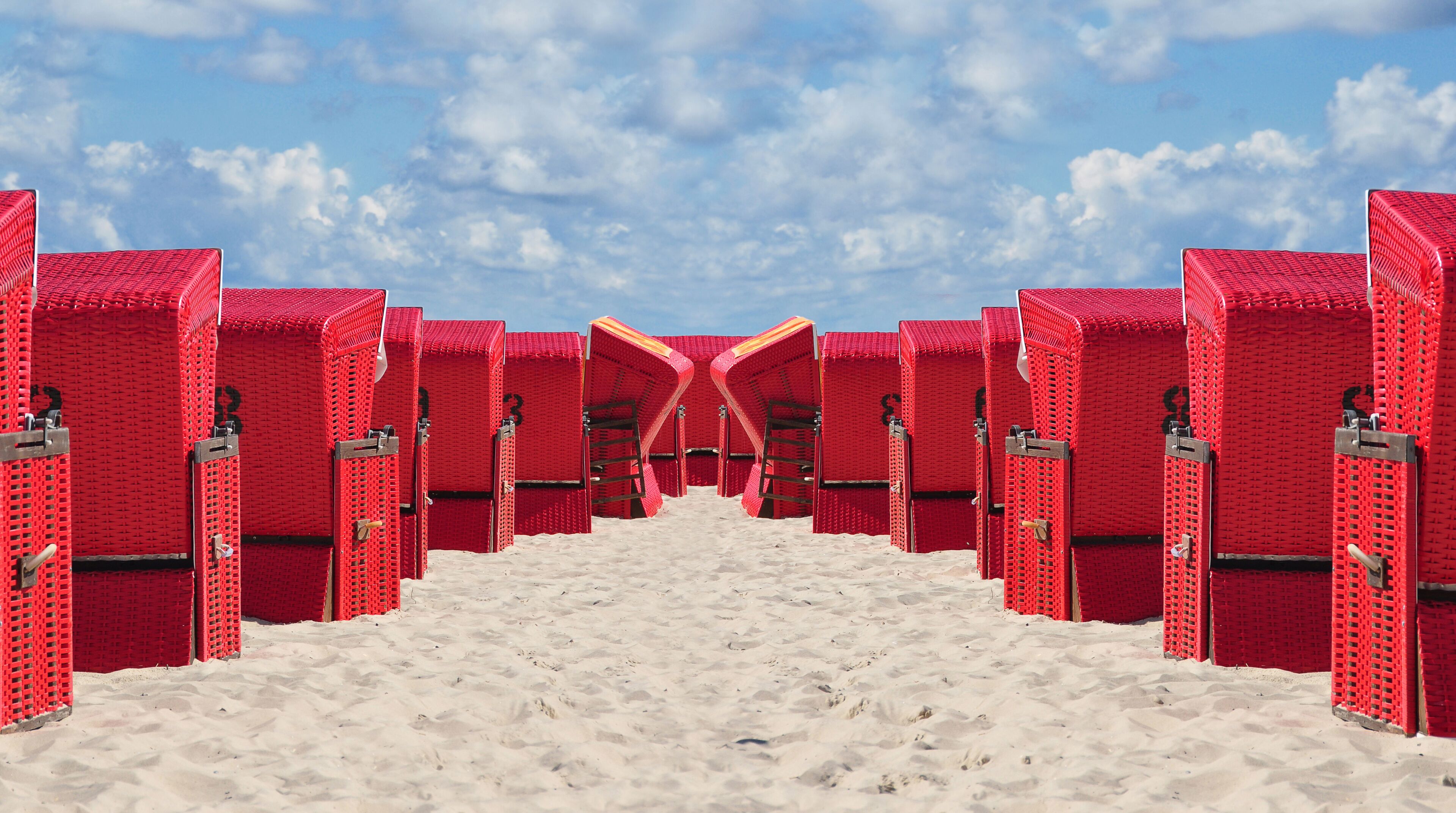 A symmetrical arrangement of red beach huts with a sandy path leading to a clear blue sky.