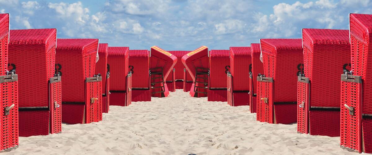 A symmetrical arrangement of red beach huts with a sandy path leading to a clear blue sky.
