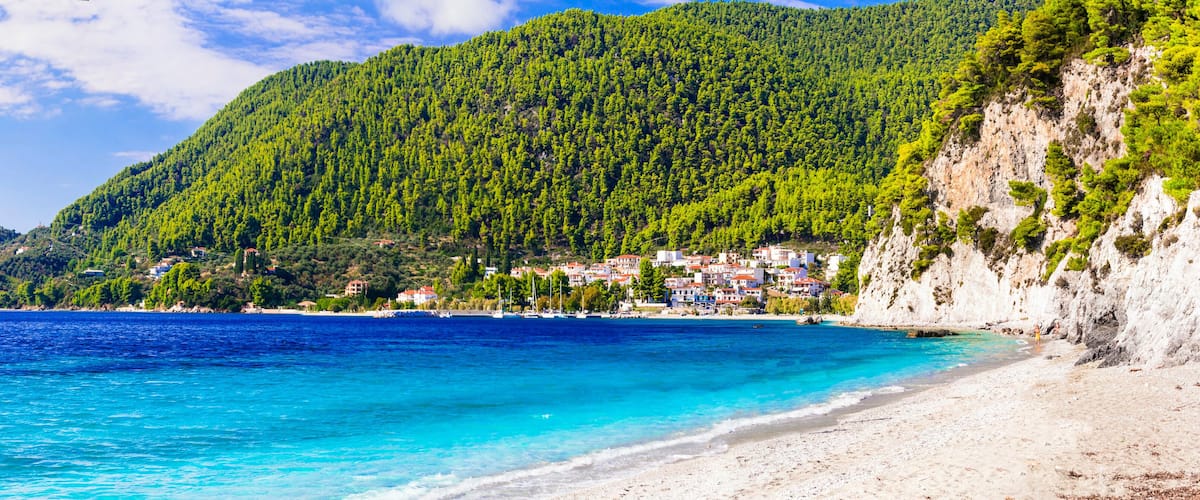 A coastal town with a beach, clear blue sea, and a forested mountain.